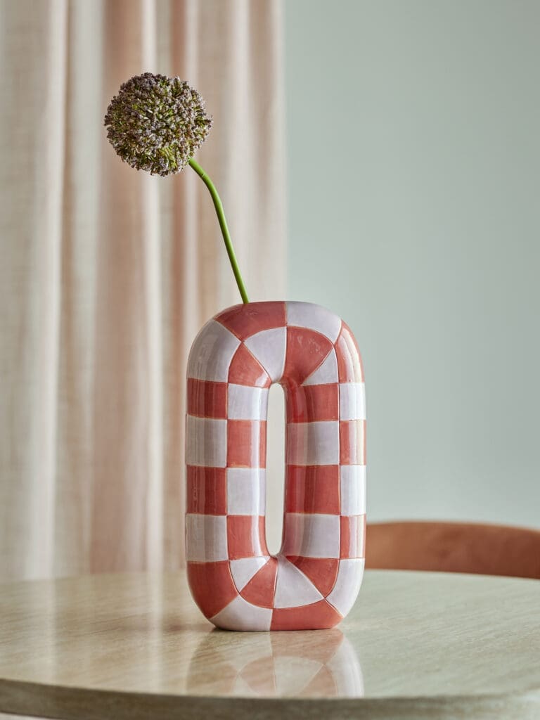 Oranje-roze geruite keramieken vaas met decoratieve bloem op een lichte houten tafel.